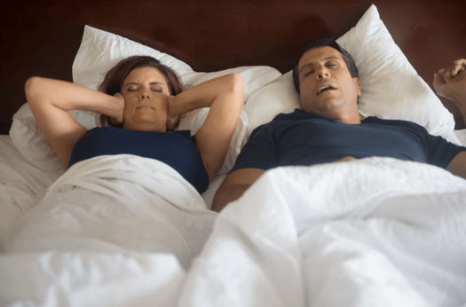 A couple lying side by side in bed with husband snoring and wife covering her ears with her hands