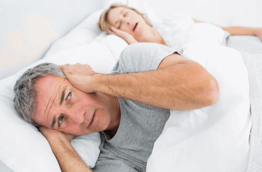 Woman asleep in bed snoring keeping up her husband who lies alongside looking unhappy about the noise and worried about her health